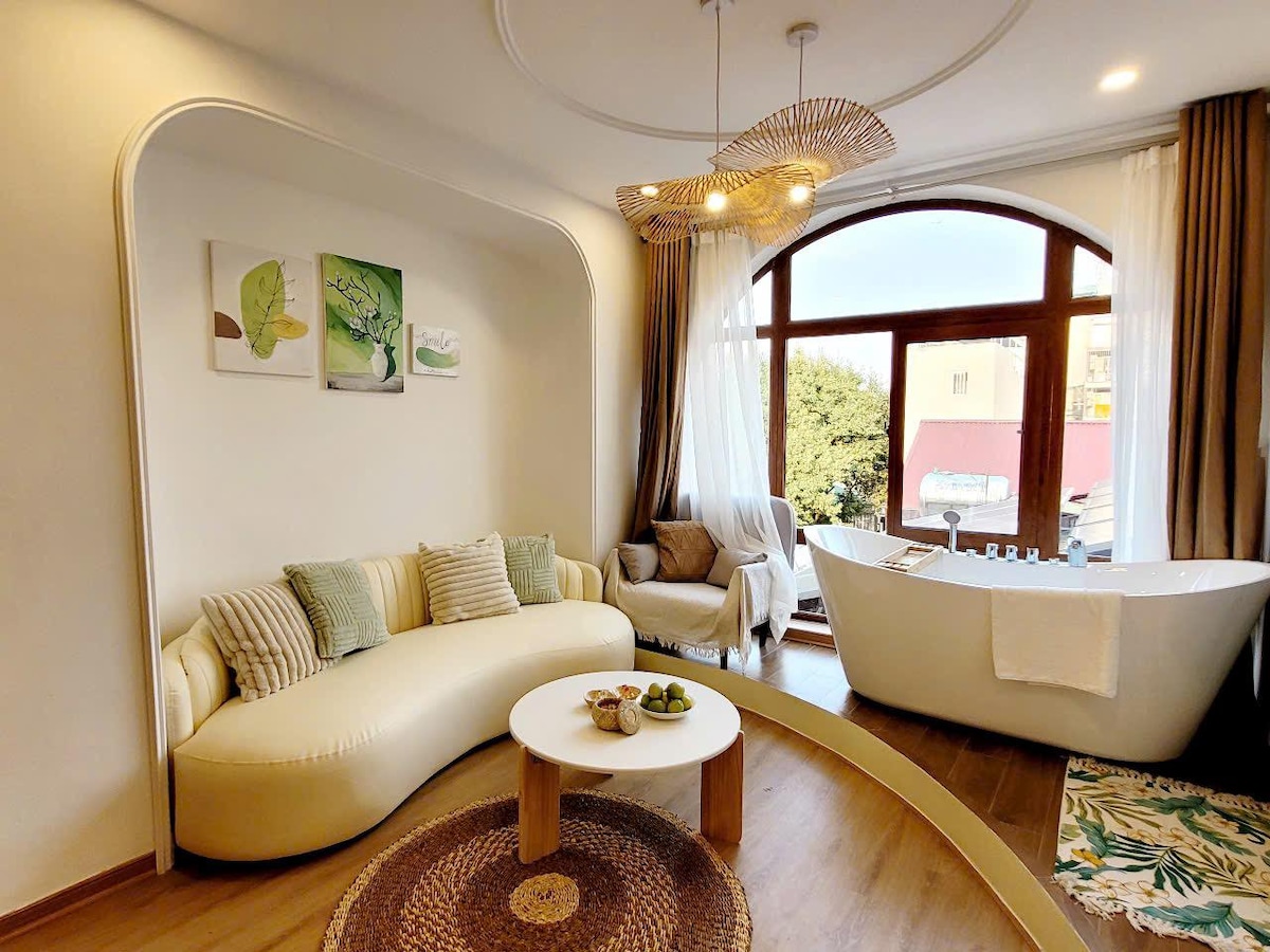 A modern living area features a curved, white sofa adorned with green and beige cushions. A round coffee table sits in front, surrounded by a woven rug. Natural light floods in through a large arched window, highlighting a luxurious bathtub nearby, complemented by artistic wall decorations.
