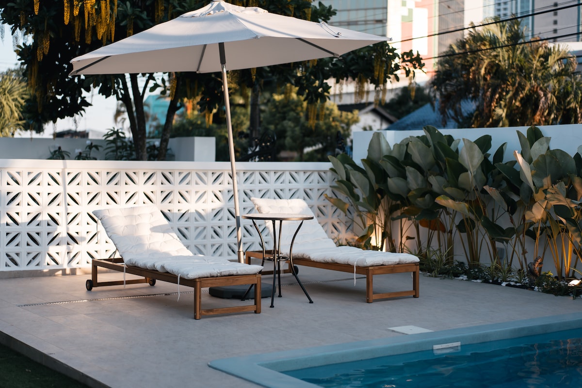 Two wooden lounge chairs with white cushions are positioned beside a dipping pool, accompanied by a small table. An umbrella provides shade, while vibrant green plants surround the area, contributing to a peaceful outdoor setting.