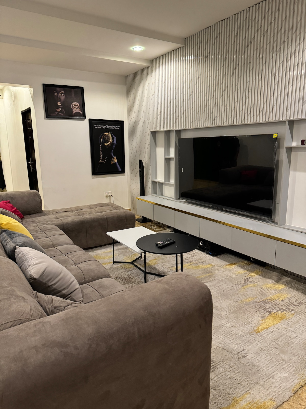 A modern living area features a spacious, gray sectional sofa adorned with colorful pillows. A sleek, low coffee table sits in front, and a flat-screen television is mounted on the wall, complemented by elegant shelving beneath. Framed artwork adds character to the space.