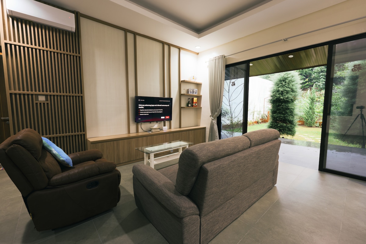 A living area features a cozy seating arrangement with a brown armchair and a light grey sofa facing a wall-mounted television. Glass doors open to a garden, allowing natural light to fill the space. Simple decor enhances the calming atmosphere.