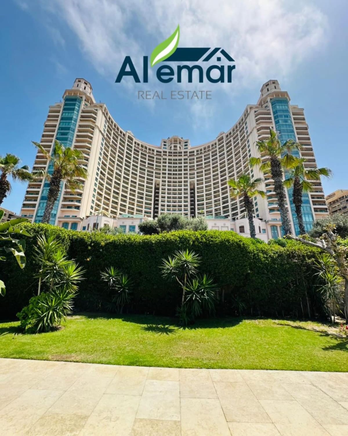 The image captures a modern high-rise building set amidst lush greenery and palm trees. The structure features multiple balconies and large windows, reflecting a bright blue sky. A neatly landscaped area is visible in the foreground, adding to the fresh atmosphere of the environment.