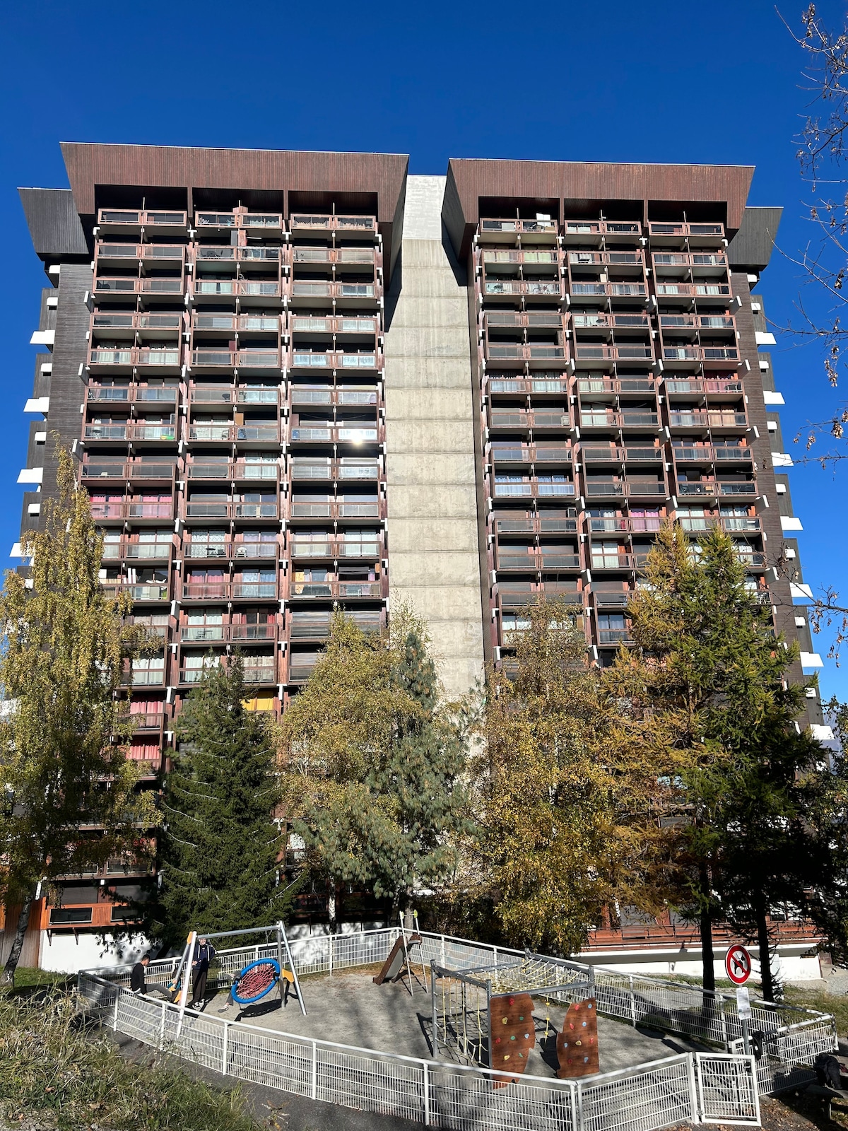 The modern building features a structured facade with multiple balconies, surrounded by trees and greenery. A playground area with climbing structures is visible in the foreground, offering recreational space. Bright blue skies provide a contrasting backdrop to the architectural design.