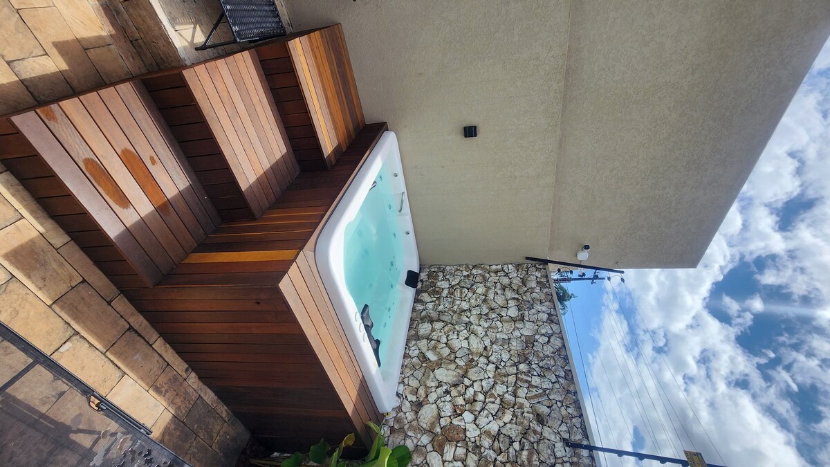 A sleek outdoor jacuzzi is positioned against a textured stone wall, surrounded by wooden decking. The serene water reflects the sky, enhancing the tranquil setting. Natural light filters in from above, creating a relaxing ambiance for guests.