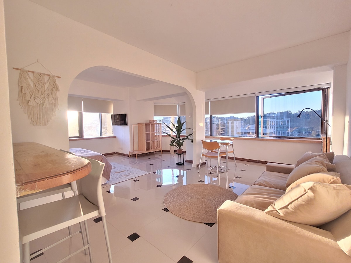A spacious living area is illuminated by natural light streaming through large windows. A comfortable sofa is positioned near a round rug, while a dining table for four is visible. Light-colored cabinets and a decorative plant enhance the open layout.