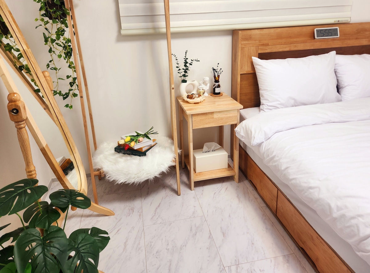 A serene bedroom scene features a spacious bed with crisp white linens. A wooden bedside table is topped with decorative items, while a soft, round rug complements the floor. A tall mirror and potted plant add warmth to the bright, inviting space.
