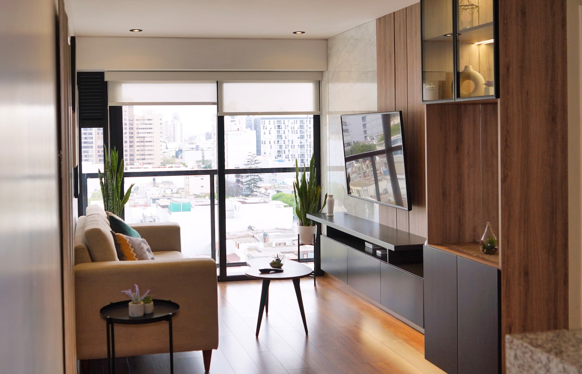 A modern living area is shown, featuring a comfortable sofa and a minimalist coffee table. Large windows allow ample natural light and showcase views of the city skyline. Wood panels adorn the walls, creating a warm ambiance, while potted plants add a touch of greenery.