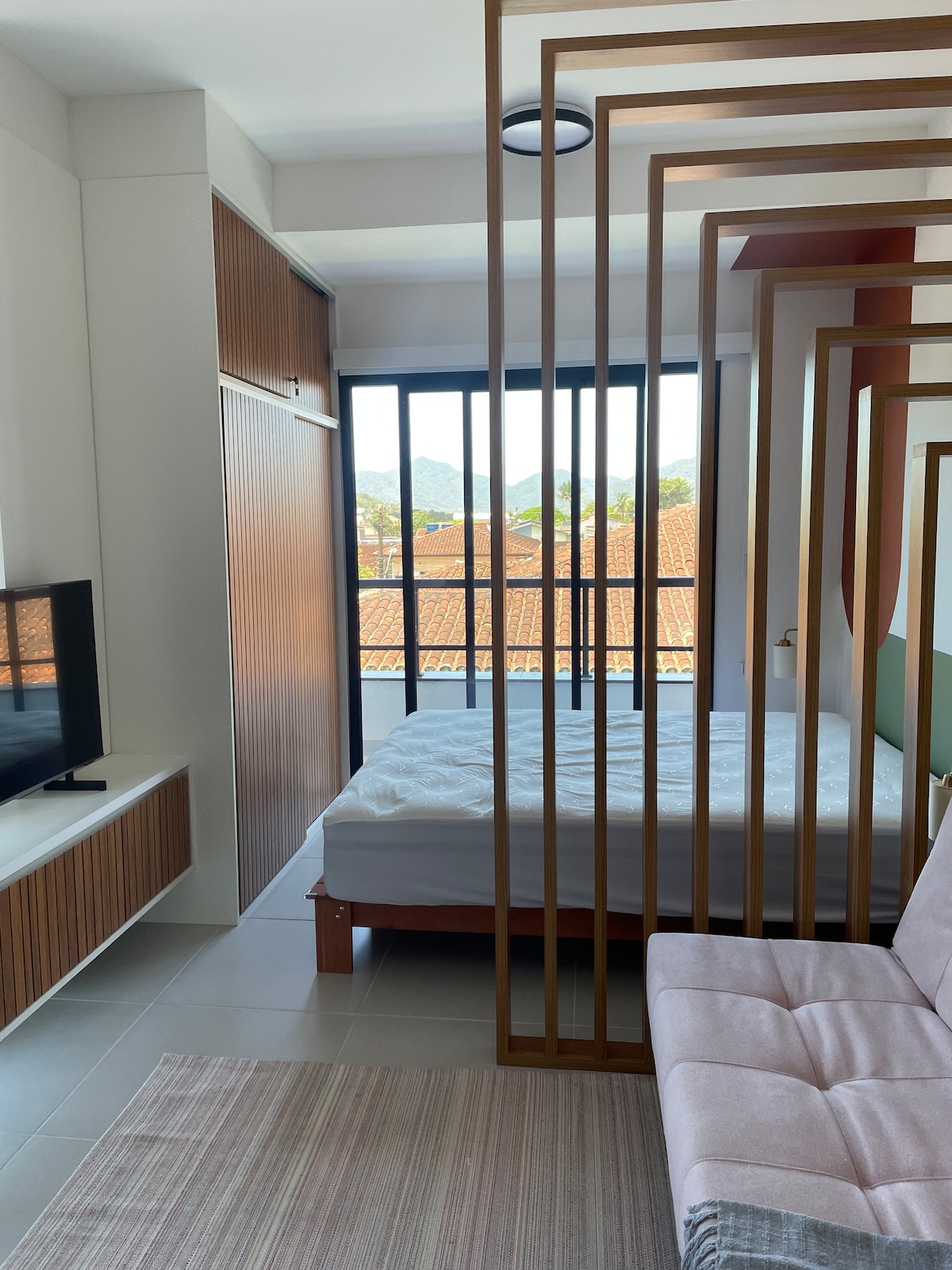 A contemporary loft interior features a defined sleeping area with a spacious bed, separated by a wooden screen. A light sofa is positioned alongside a television unit, with natural light illuminating the room through large windows overlooking tiled rooftops and distant mountains.