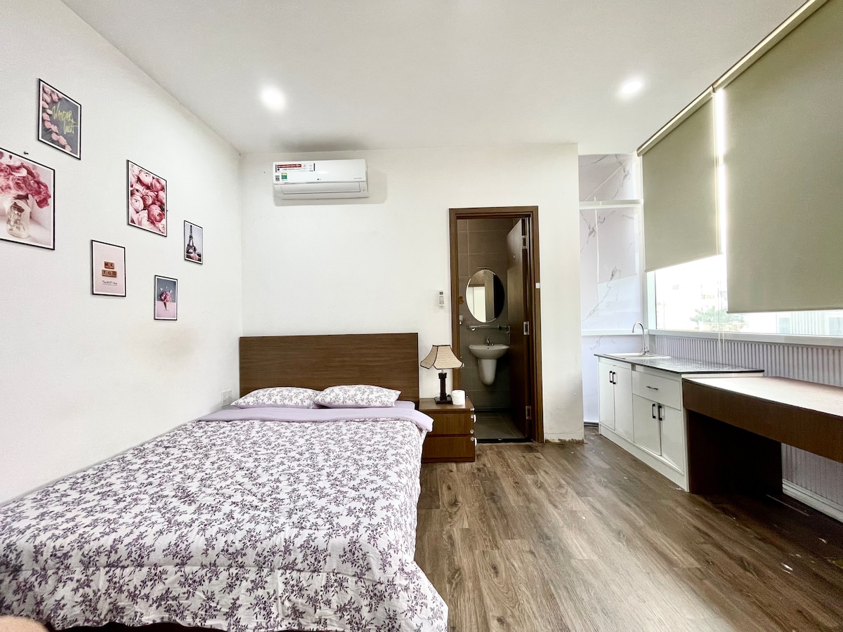 A serene sleeping area features a wooden bed adorned with a floral-patterned bedspread. A framed artwork decorates the wall. A desk area is present, along with a lamp. A door leads to a bathroom, and large windows allow natural light to brighten the space.