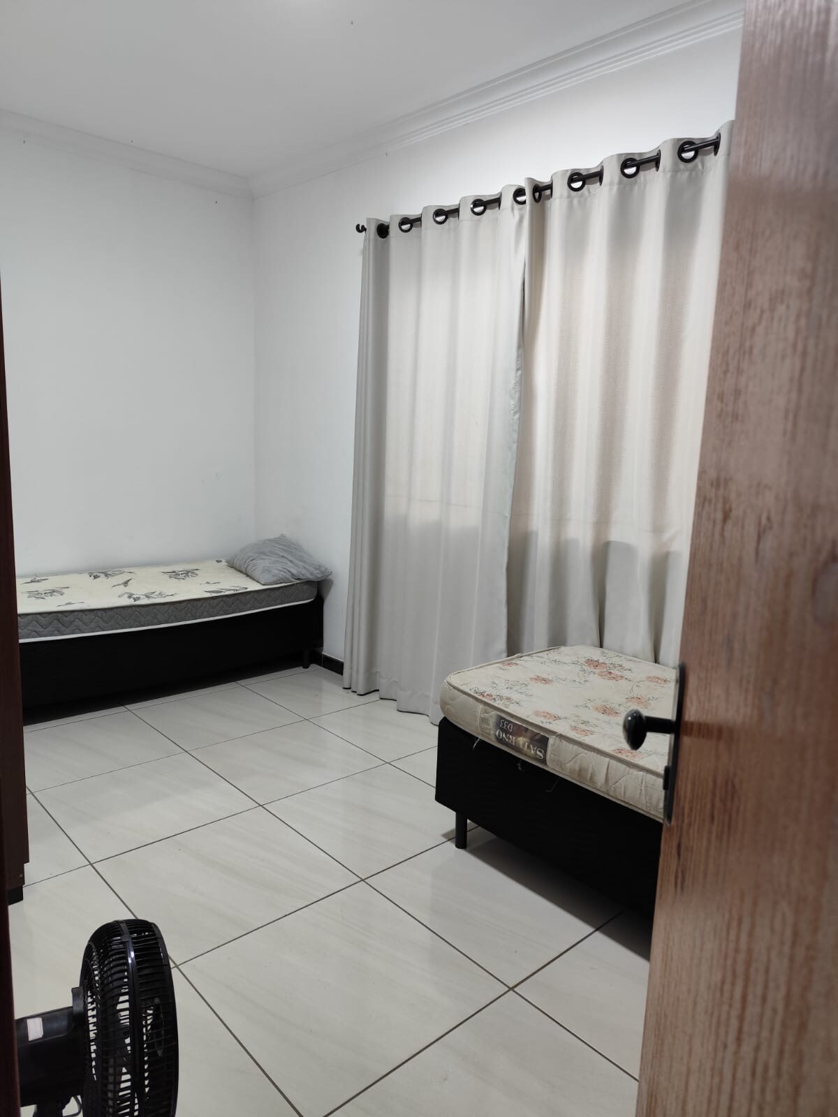 A simple guest room is displayed, featuring two beds with neutral-toned bedding. Light-colored curtains filter natural light, while the floor is tiled in a uniform pattern. A small fan is positioned near the door, and a wooden door frame adds a warm touch.