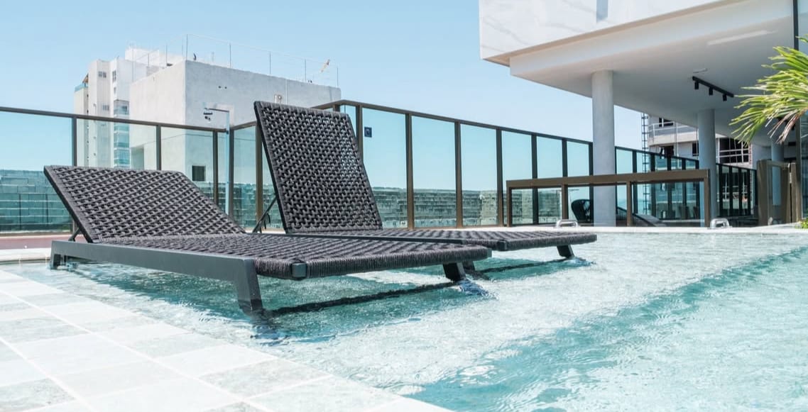 A sunlit pool area features two lounge chairs positioned at the water's edge. The clear water reflects the bright blue sky, providing a serene atmosphere. Surrounding the pool is a glass railing, enhancing the openness of the space.
