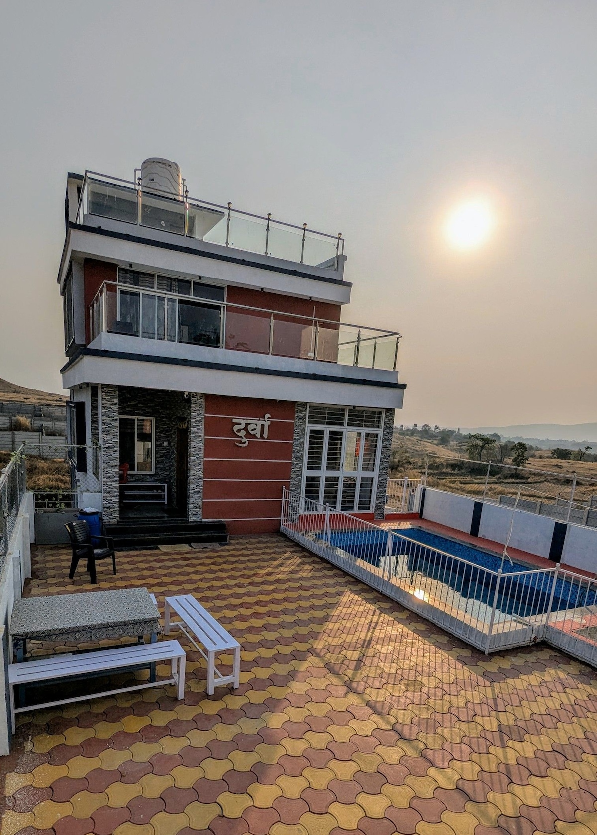 A three-story structure is presented with large glass windows and a spacious balcony. The exterior showcases a blend of brick and modern materials. A rectangular pool is situated at the forefront, surrounded by a tiled area. The sun is setting in the background, creating a serene ambiance.