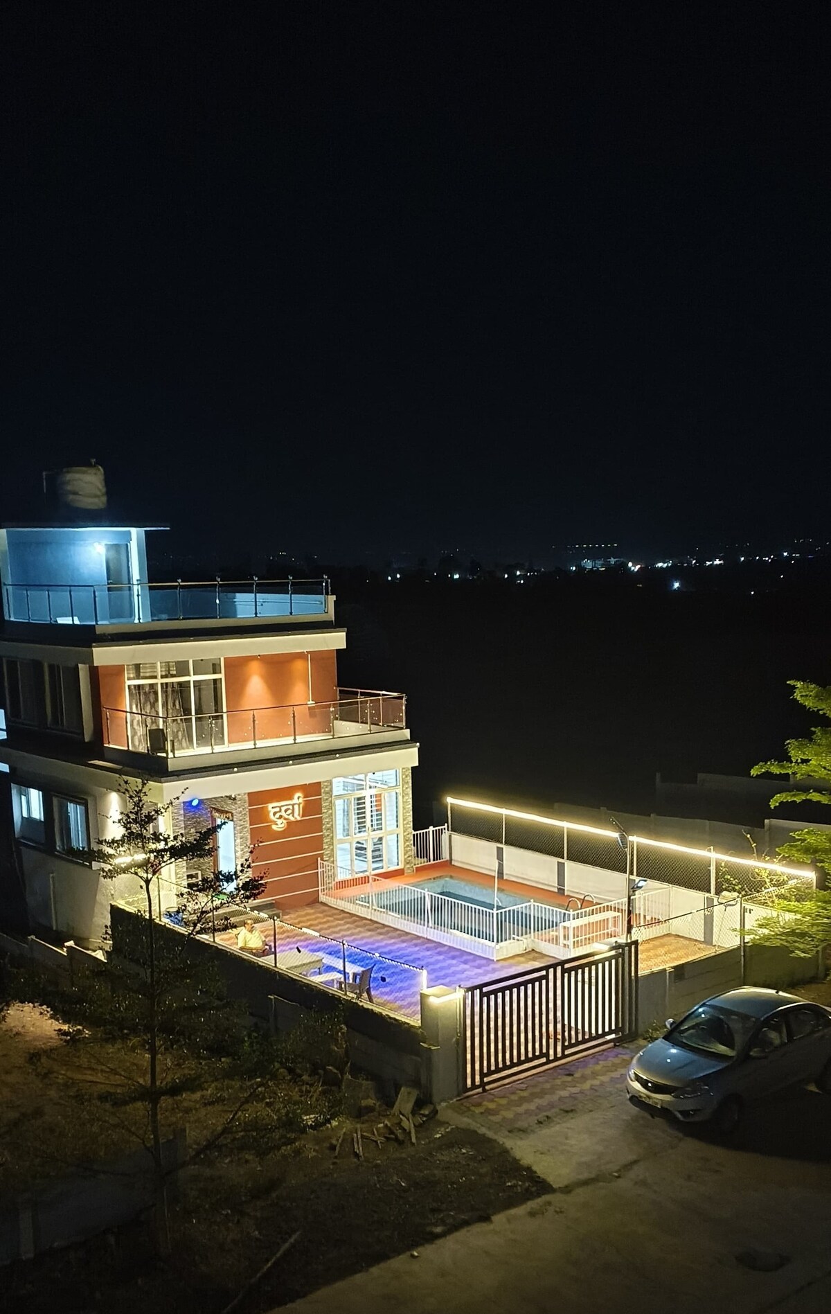 A modern three-story house is shown at night, featuring illuminated exterior lighting that highlights its design. A swimming pool is visible within a fenced area, while city lights can be seen in the background, providing a serene nighttime ambiance.