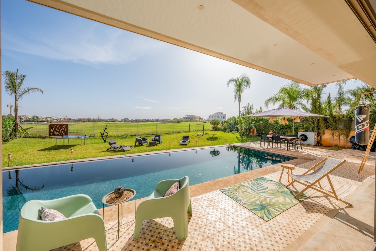 A spacious terrace overlooks a tranquil swimming pool, framed by palm trees and a lush green golf course. Lounge chairs are arranged beside the pool, creating a relaxing outdoor space. A dining area is set under a canopy, inviting meals with a view.