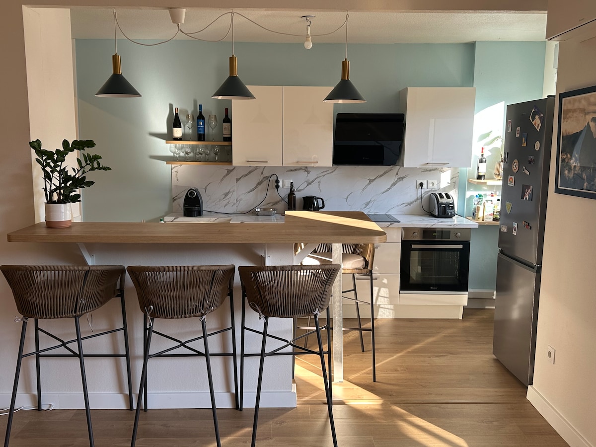 A modern kitchen space is highlighted by a wooden bar with four high-backed chairs. Sleek white cabinetry complements a decorative glass shelf with various bottles. Stainless steel appliances, including an oven and refrigerator, add functionality, while soft natural light illuminates the room.