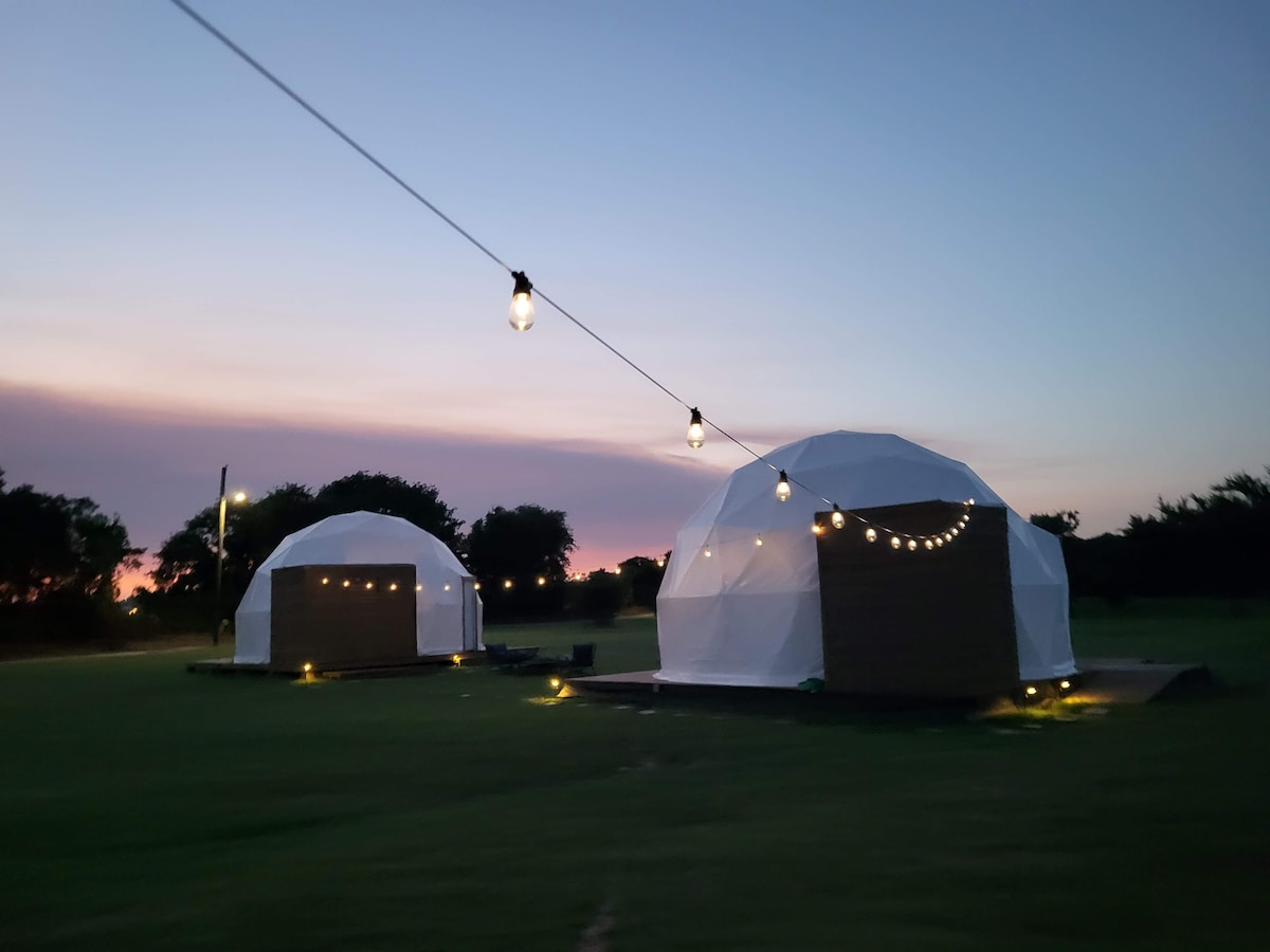 Two spacious domes are illuminated by string lights at twilight, set against a vibrant sunset sky. Green grass surrounds the domes, creating an inviting outdoor space for gatherings.