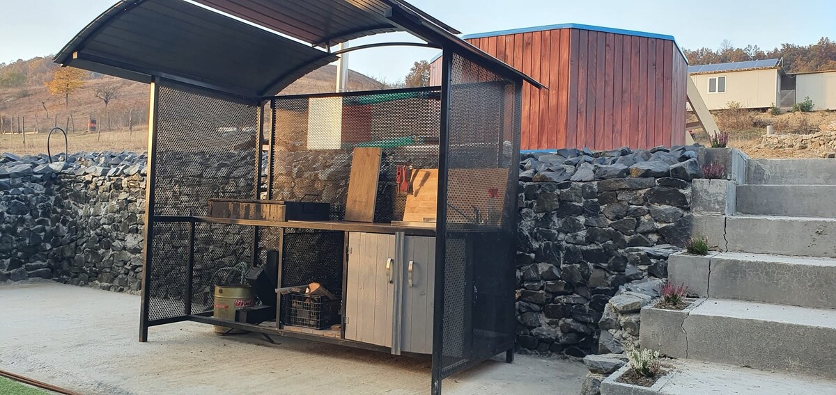 An outdoor cooking area is featured, constructed with mesh and metal under a protective roof. The kitchen space includes a work surface, storage cabinet, and various utensils. The backdrop consists of a stone wall, enhancing the rustic atmosphere. Stairs lead up to an adjacent building.