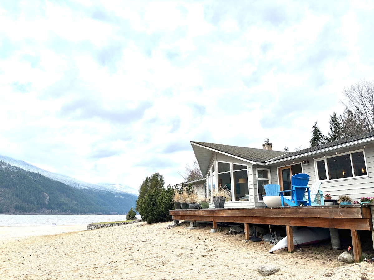A lakefront view of the beach house is presented, showing a wooden deck extending over sandy beach. The house features large windows and a modern design, with colorful outdoor chairs placed on the deck. Surrounding mountains and serene water can be seen in the background.
