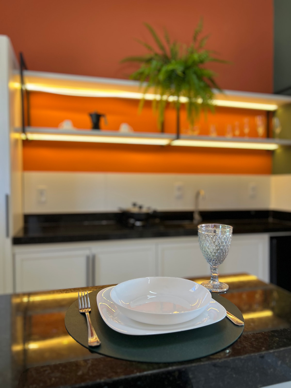 A modern kitchen is highlighted by a sleek black countertop and vibrant orange wall. A neatly set table features white plates and polished cutlery, with a decorative glass standing beside it. Shelves above contain various dinnerware, accented by a green plant for a touch of freshness.