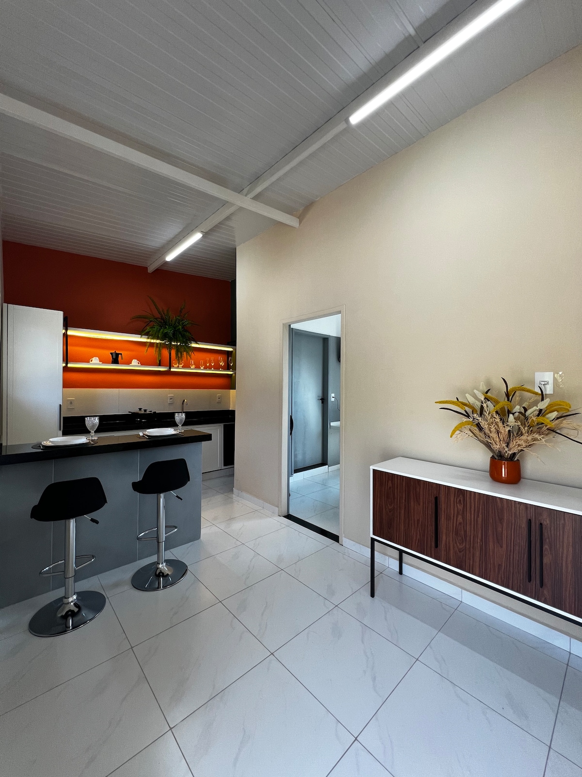 The kitchen features modern appliances against a backdrop of orange and earthy tones. A sleek countertop is paired with two black bar stools. Natural light illuminates the space, highlighting the decorative plant and a stylish cabinet with minimalist decor.
