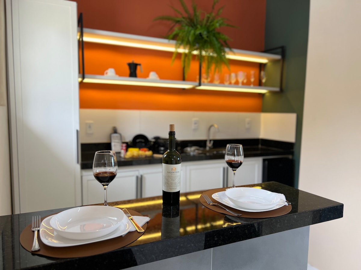 A sleek kitchen area features a dark countertop with two elegant plates and wine glasses set for a meal. A bottle of wine is prominently displayed. The background highlights warm-colored wall accents and organized shelves with glassware, creating an inviting space.