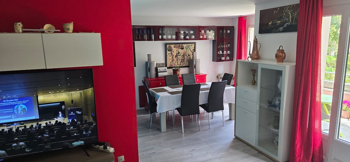 A vibrant living area is displayed, featuring a large television and a sleek white cabinet. A dining table with six chairs is centrally positioned against a bold red accent wall. Sunlight filters through windows adorned with light red curtains, illuminating the space.