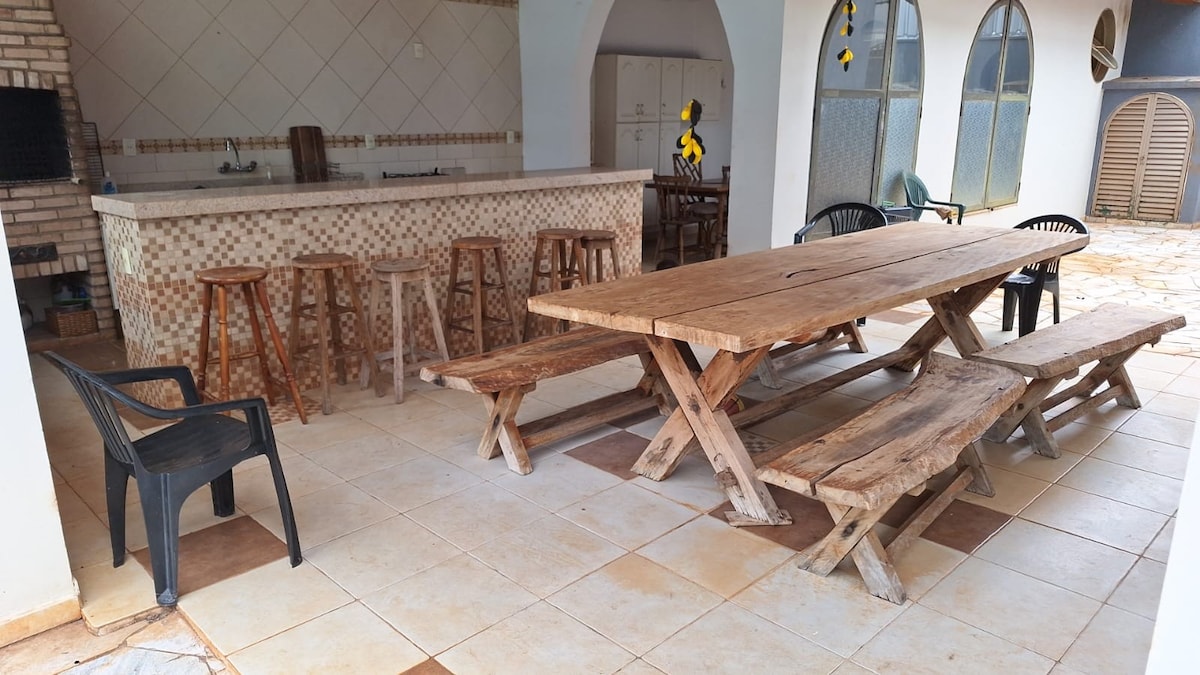 A spacious outdoor dining area features a long wooden table surrounded by benches. Several black chairs are placed nearby. Behind, a kitchen counter with bar stools is visible, accented by a light tile pattern. Arched windows allow natural light into the space.