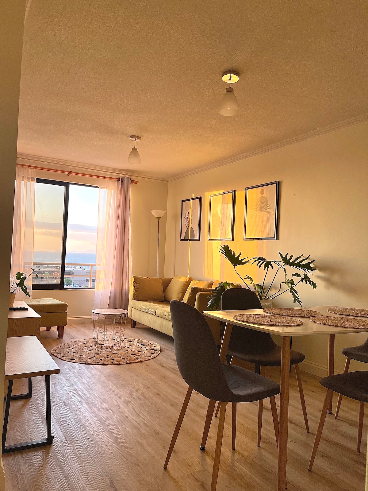 A well-lit living area features a comfortable sofa and a dining table with four chairs. Large windows allow for natural light and offer a view of the sunset. Decorative plants provide a touch of greenery, enhancing the inviting environment.