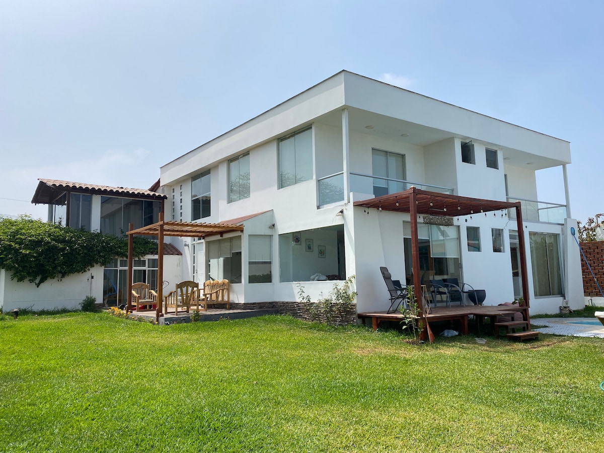 The two-story house features a modern design with large windows, offering ample natural light. A deck with seating is visible in the foreground, adjacent to expansive green lawn. The surrounding area is enhanced by trees and plants, creating a serene outdoor environment.