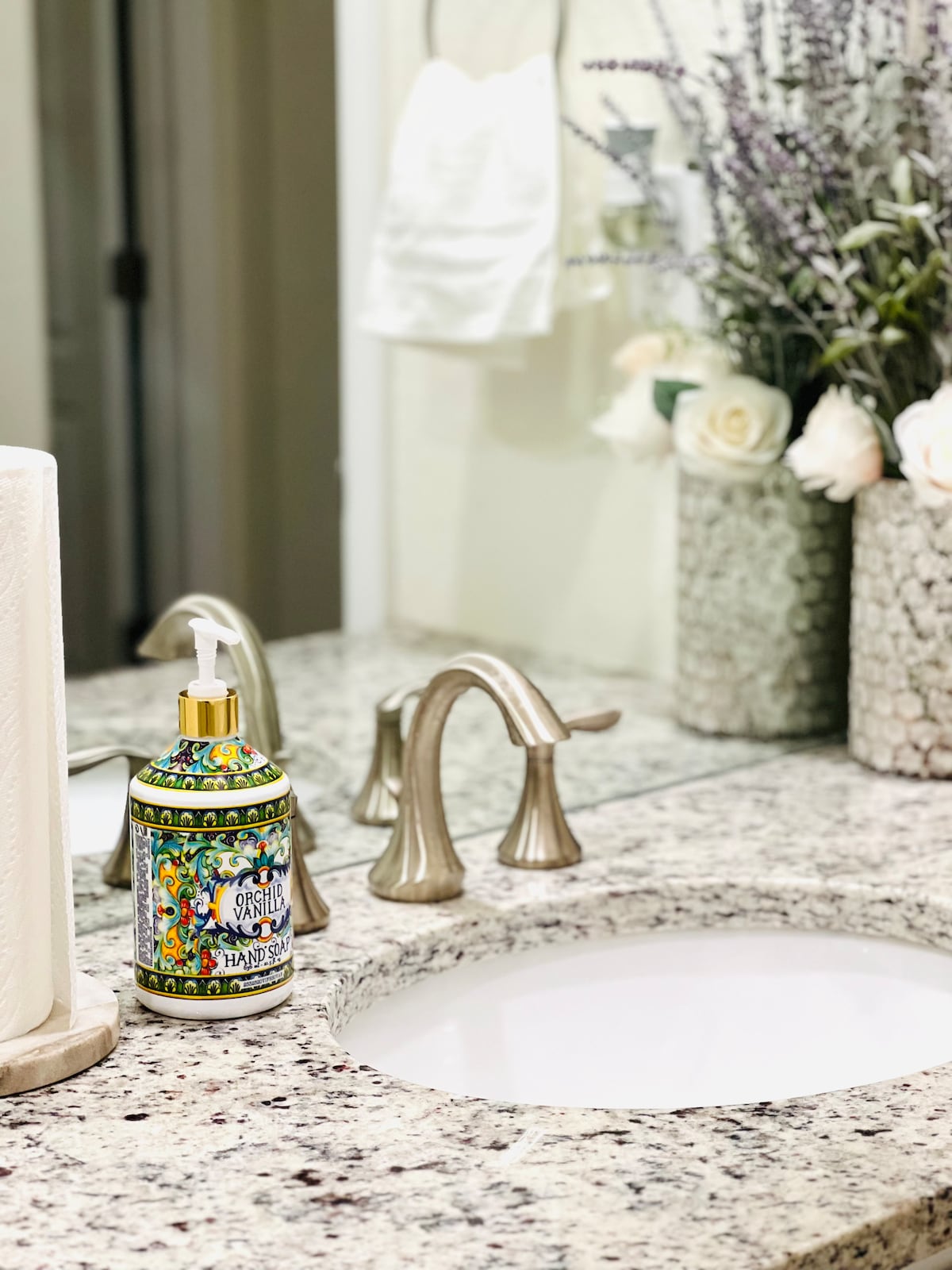 The bathroom features a granite countertop with dual sinks and elegant fixtures. A decorative soap dispenser, patterned with vibrant colors, is placed nearby. Fresh flowers in textured containers add a touch of greenery, enhancing the calming ambiance of the space.