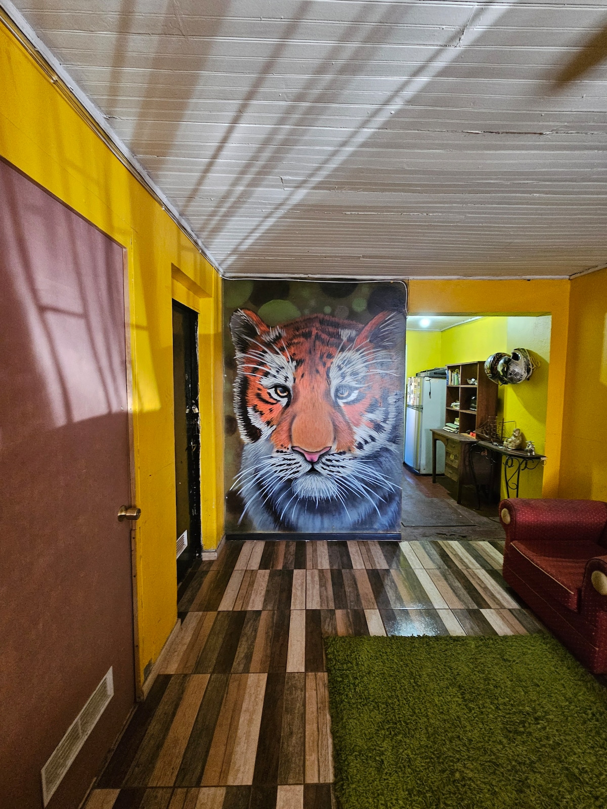 A hallway features a striking mural of a tiger on one wall, surrounded by vibrant yellow paint. The floor is covered in alternating wood-patterned tiles, with a plush green carpet underfoot. A cozy red seating area is visible in the background, creating a unique and inviting space.