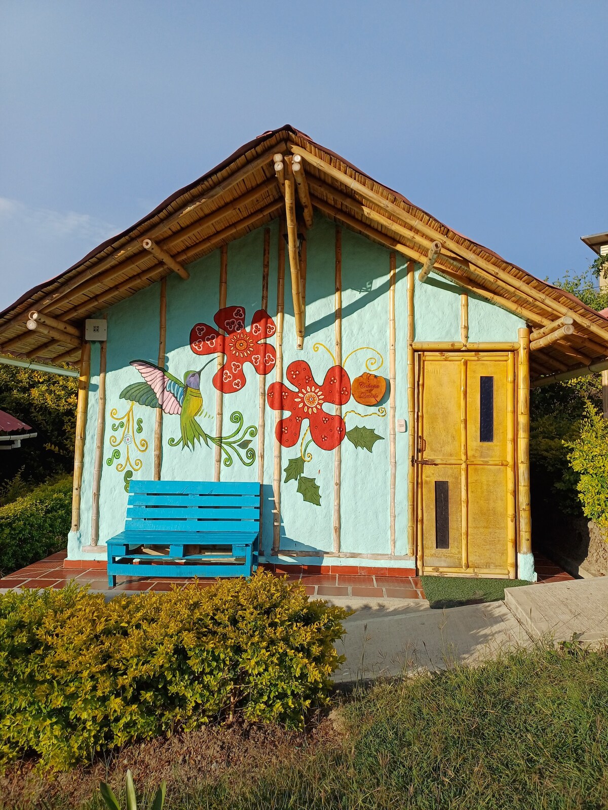 A cabin exterior showcases a charming mural of colorful flowers against a light blue wall. A wooden door with a golden hue complements the design. A bright blue bench is positioned on a tiled patio, surrounded by lush greenery and shrubs.