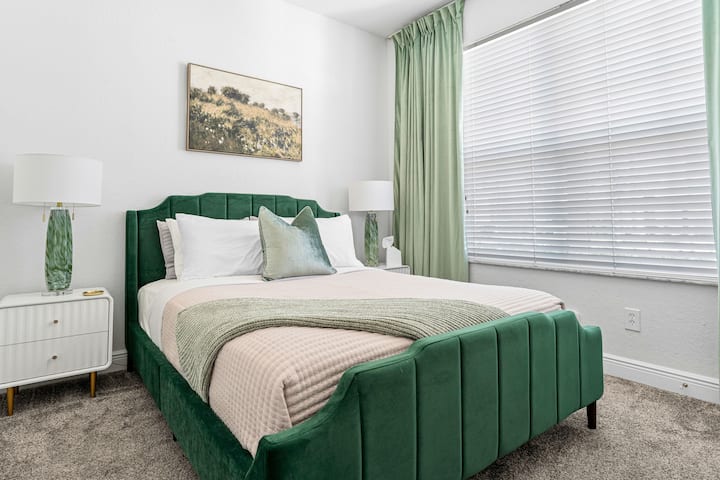This elegant bedroom features a plush queen-size bed with stylish green upholstered frame and matching curtains. A smart TV provides entertainment, while large windows fill the space with natural light, creating a serene and comfortable retreat.