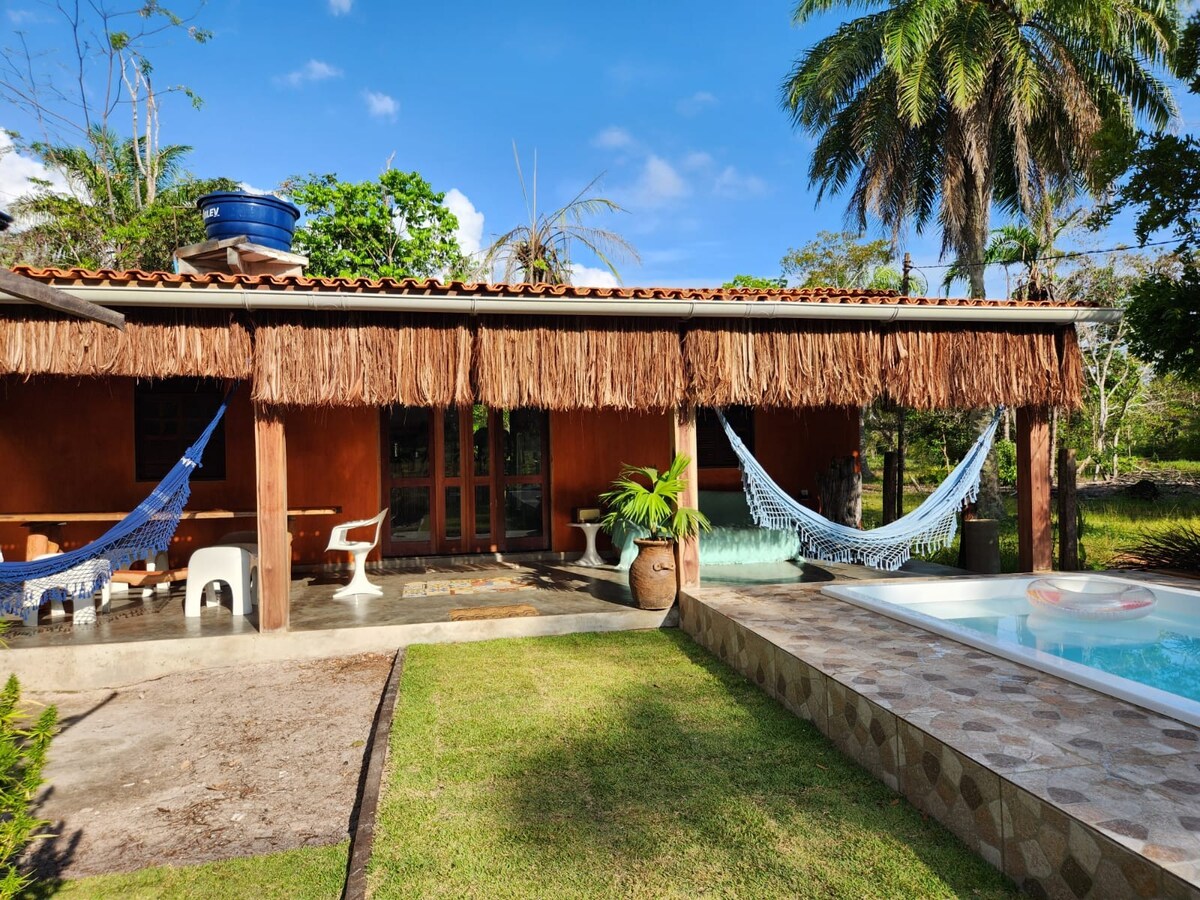 An outdoor area displays a thatched roof providing shade over a spacious patio. Comfortable hammocks are suspended for relaxation, accompanied by a small table and chairs. A pool is visible nearby, with lush greenery enhancing the tranquil atmosphere.