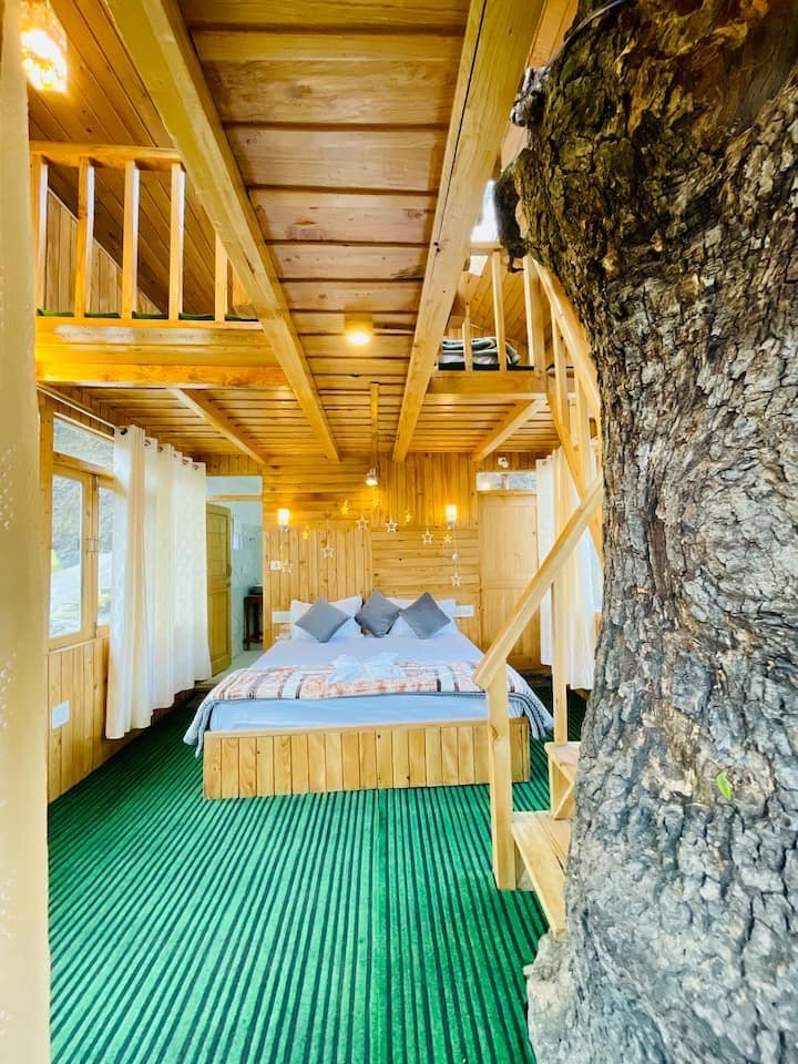 A cozy treehouse interior features wooden walls and flooring, creating a warm atmosphere. A spacious bed is centered in the room, flanked by decorative pillows. A staircase leads to an upper loft, with soft lighting illuminating the space. A natural tree trunk enhances the unique character.