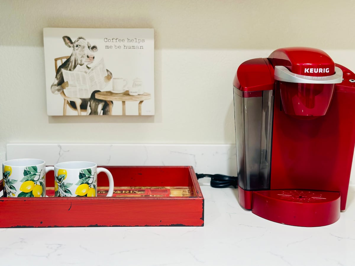 A vibrant red Keurig coffee maker is positioned on a white countertop, accompanied by two lemon-patterned mugs. A rustic red tray holds a magazine and a decorative sign that reads 'Coffee helps me be human,' enhancing the kitchen's cozy functionality.