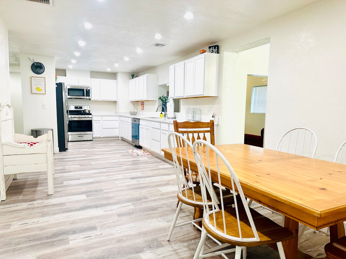A spacious kitchen and dining area is visible, featuring white cabinetry and stainless steel appliances. A large wooden dining table with six chairs is positioned prominently. Bright overhead lighting enhances the room's functionality, while a glimpse of a separate living area can be seen through an open doorway.