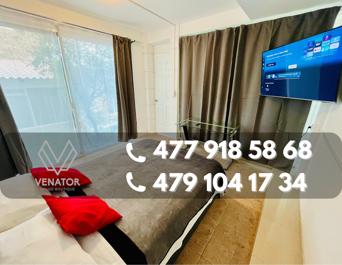 A comfortable sleeping area is presented with a spacious bed dressed in neutral bedding, complemented by red accent pillows. A flat-screen TV is mounted on the wall, and large windows covered by dark curtains allow soft light to filter in.
