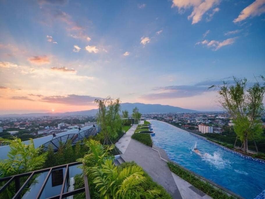The image captures a panoramic view of a rooftop swimming pool that stretches along the edge of a building. Lush greenery lines the pool area, and a vibrant sunset is visible over the distant mountains and cityscape.
