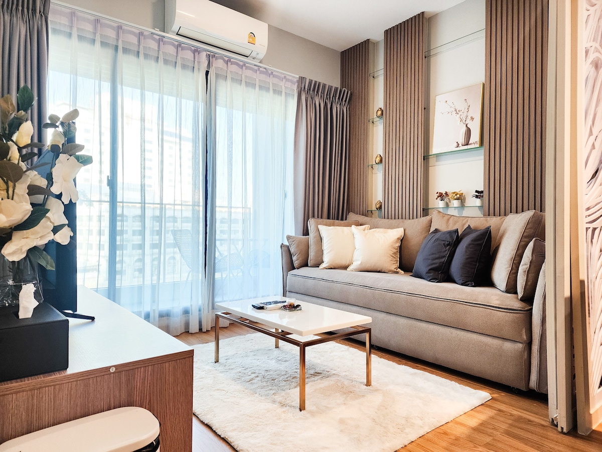 A comfortable living area is visible, featuring a spacious grey sofa adorned with dark cushions. Natural light illuminates the space through sheer curtains, and a coffee table sits atop a soft rug. A small balcony is accessible through glass doors, enhancing the overall openness.