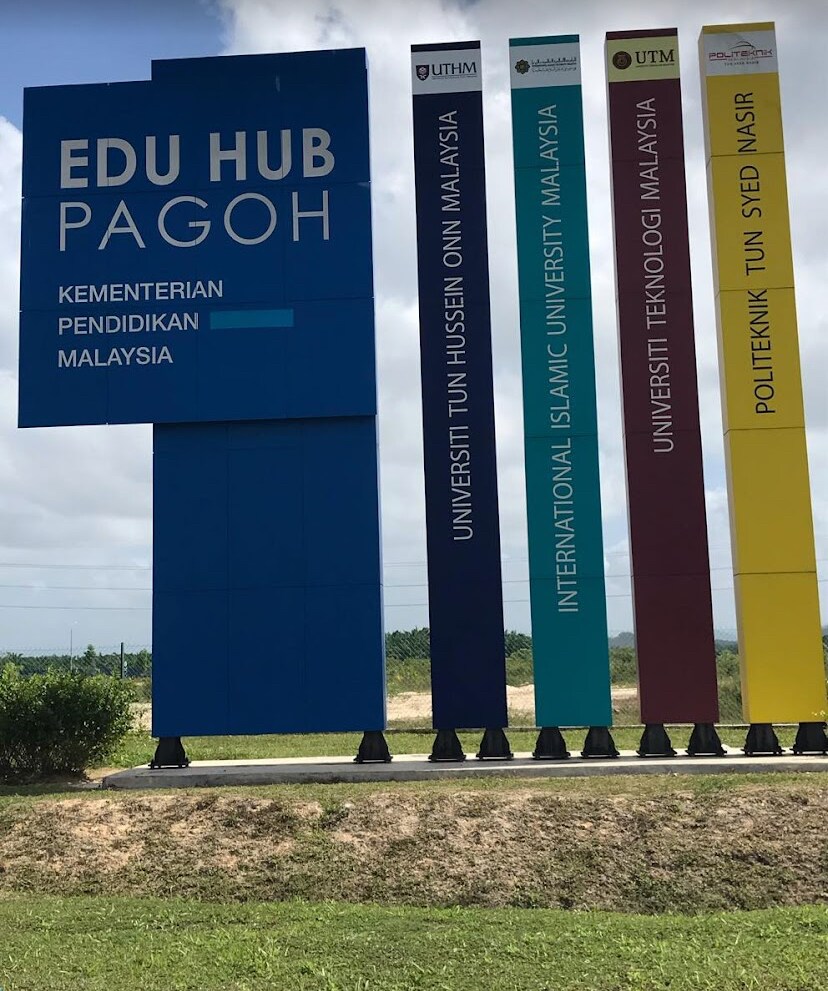 A large, colorful sign displays the names of various educational institutions, including Universiti Tun Hussein Onn Malaysia and Universiti Teknologi Malaysia. The bold letters are set against a clear sky, with greenery visible in the background, highlighting the hub's educational significance.