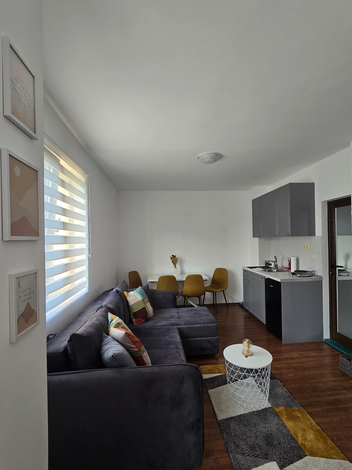 A modern living area features a dark sectional sofa with decorative pillows, complemented by a round coffee table. A dining space with four yellow chairs is positioned near the window, while a compact kitchen with sleek cabinetry and appliances occupies one corner of the room.