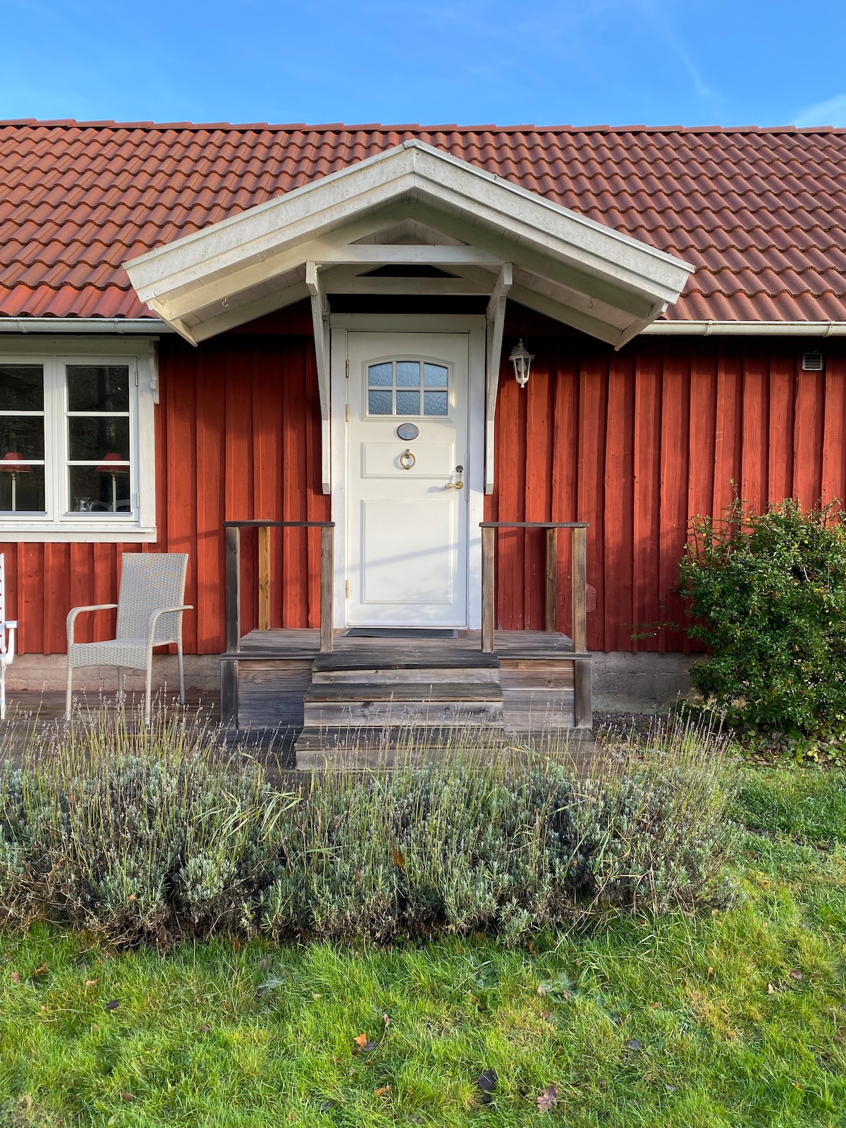 A charming entrance is featured with a white door framed by a small overhead awning. A wooden staircase leads to the door, and a single chair sits to the side. The vibrant red exterior contrasts with surrounding greenery and lavender plants, creating a welcoming approach.