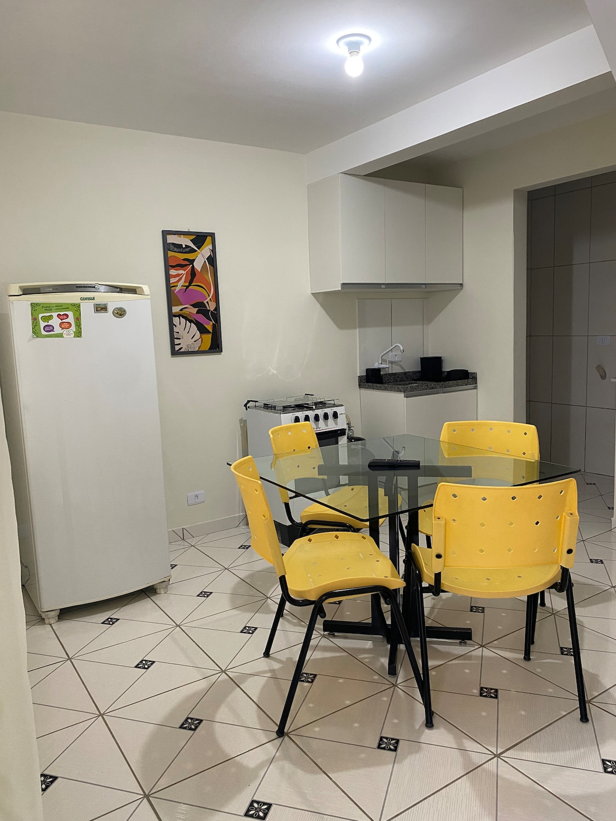 A modern kitchen area is depicted, featuring a glass dining table surrounded by four yellow chairs. A white refrigerator and additional cabinetry are visible, alongside a counter with kitchen appliances. A piece of vibrant wall art adds a touch of color to the neutral walls.