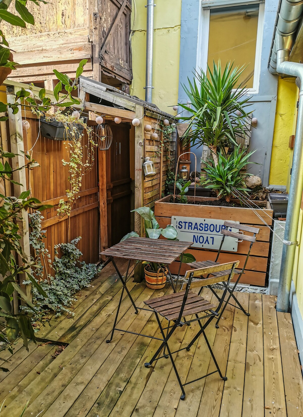 An outdoor terrace is visible, featuring a small wooden table with two black folding chairs. Lush greenery, including potted plants and climbing vines, enhances the atmosphere. Soft lighting is provided by hanging lights, creating a cozy and inviting space in the calm surroundings.