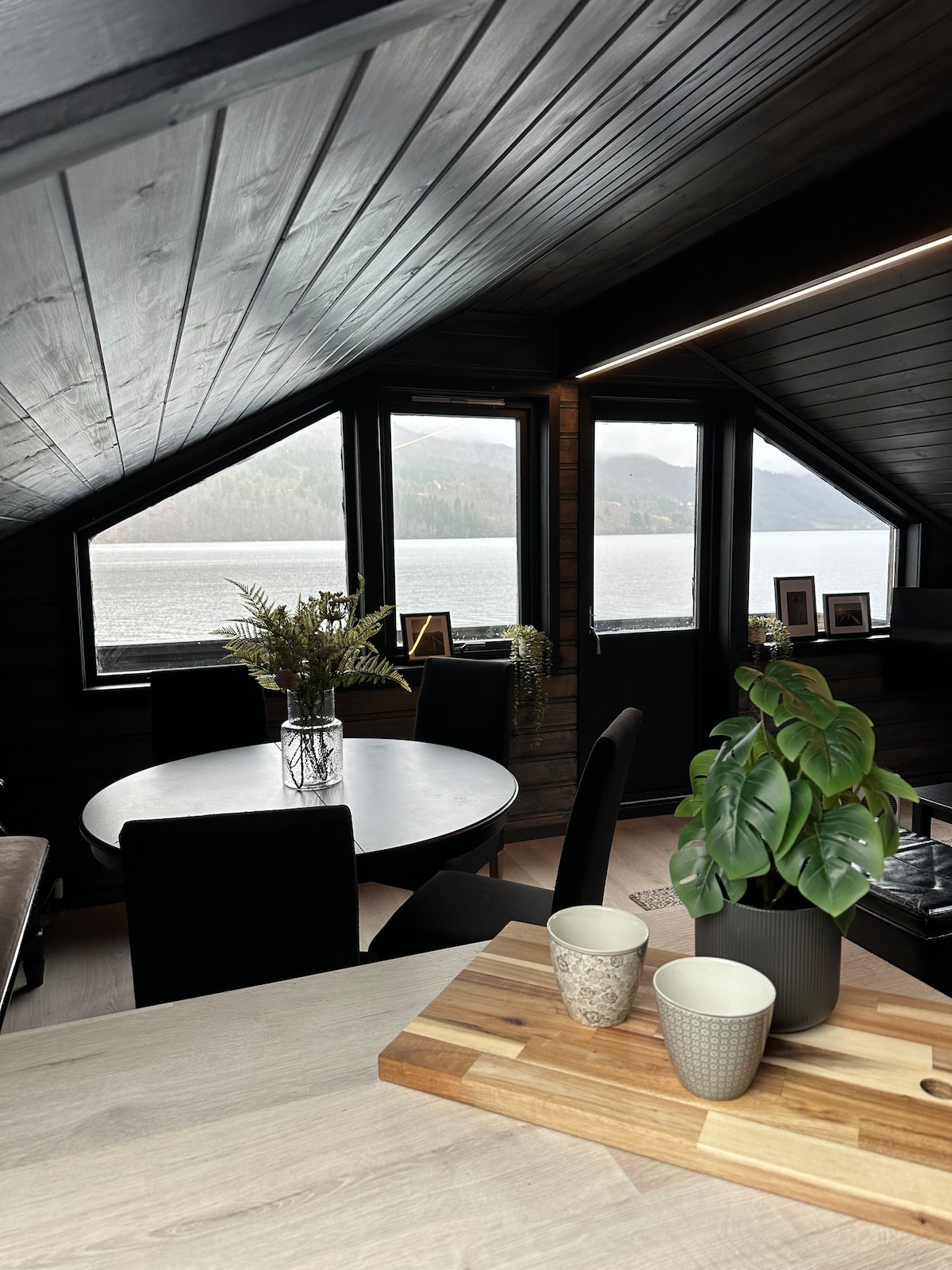 A dining area features a round table surrounded by four chairs, with potted plants adding a touch of greenery. Large windows offer a view of a serene lake and surrounding nature. Dark wood accents create a cozy atmosphere, enhanced by subtle lighting.