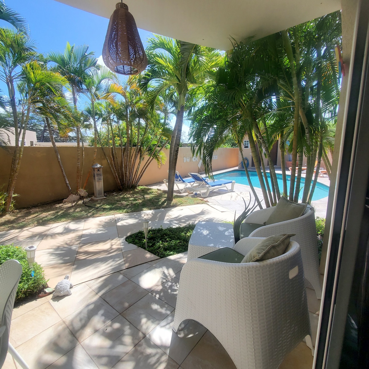 A serene outdoor patio is surrounded by lush greenery and palm trees. Comfortable white lounge chairs are positioned near a small shared pool, offering a perfect spot for relaxation. The space is designed for tranquil enjoyment, with natural light filtering through the foliage.