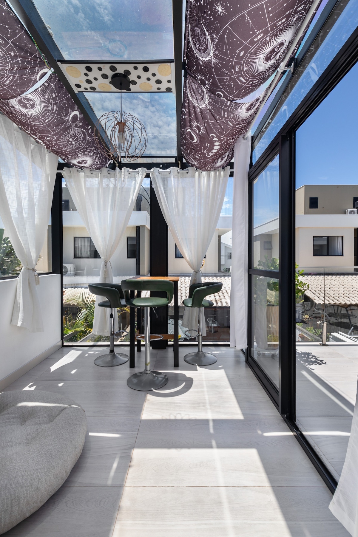 A sunlit terrace features floor-to-ceiling glass windows, allowing for ample natural light. Two high-backed chairs and a small round table create an inviting space for relaxation. Sheer white curtains frame the scene, while decorative fabric overhead adds a touch of style to the comfortable setting.