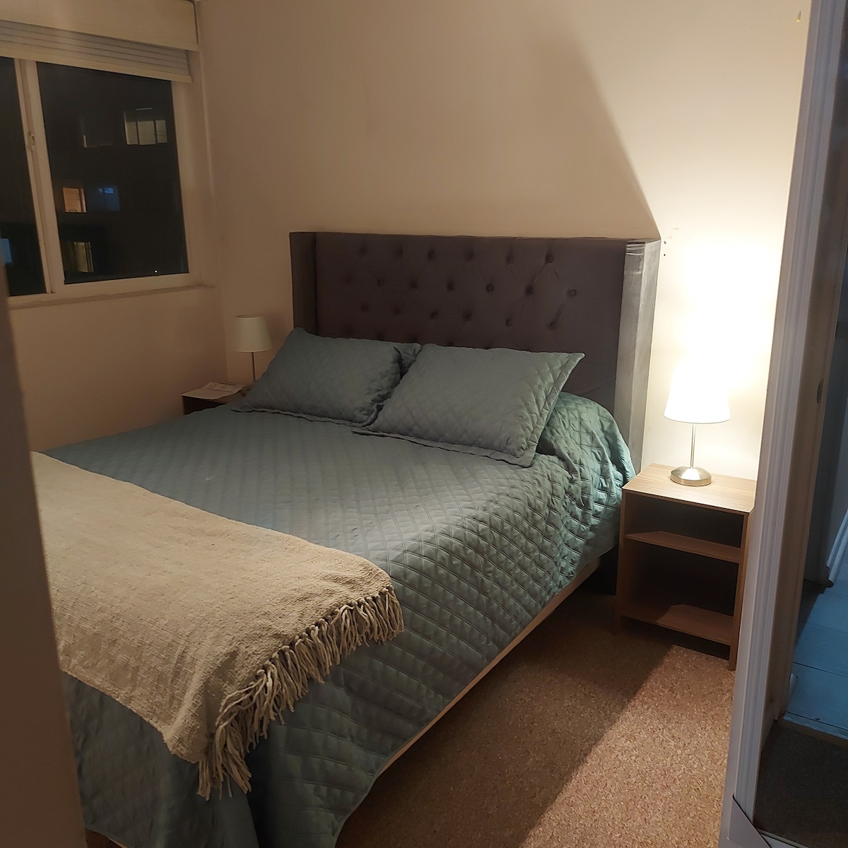 A well-lit sleeping area features a spacious bed with a patterned quilt and two pillows. Bedside tables hold lamps providing soft light. A window allows for natural light, and a neutral-toned throw lies casually across the foot of the bed.