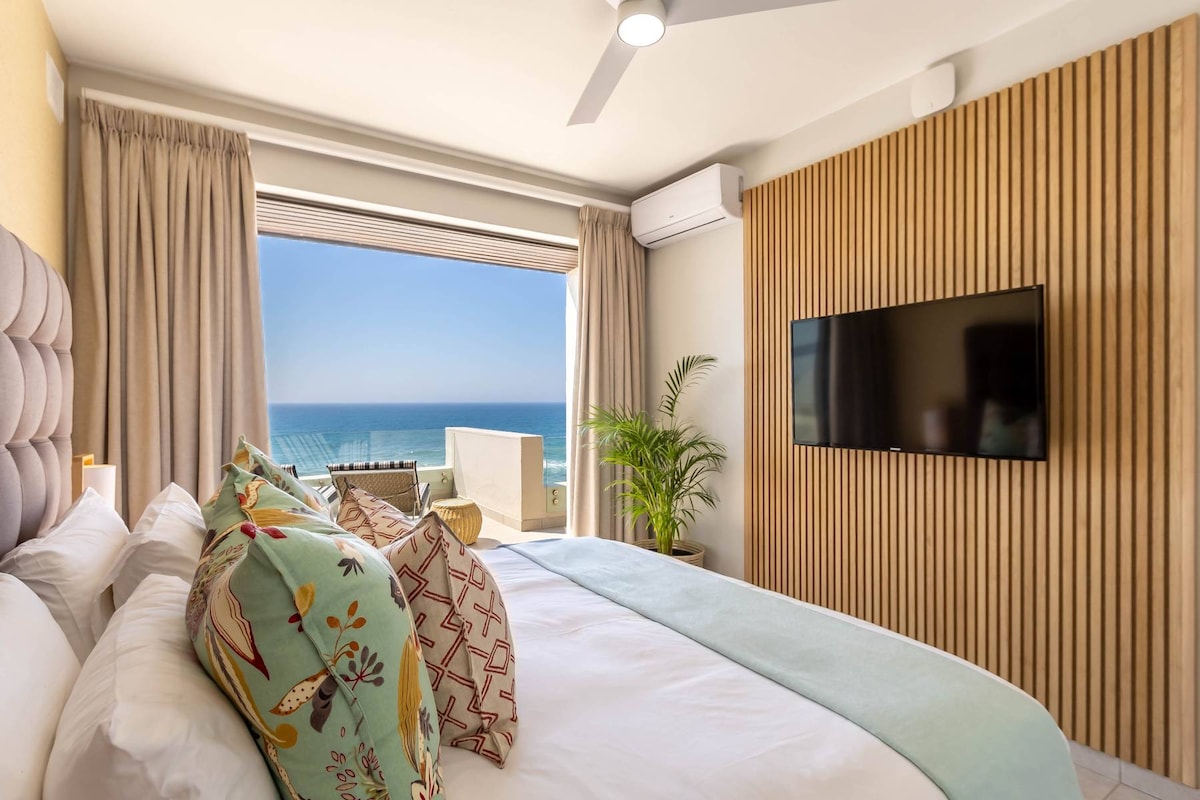 A bright and inviting bedroom features a spacious bed adorned with decorative pillows. Large windows offer elevated views of the beach and ocean, while a flat-screen television is mounted on the wooden-paneled wall. Natural light fills the room through sheer curtains.
