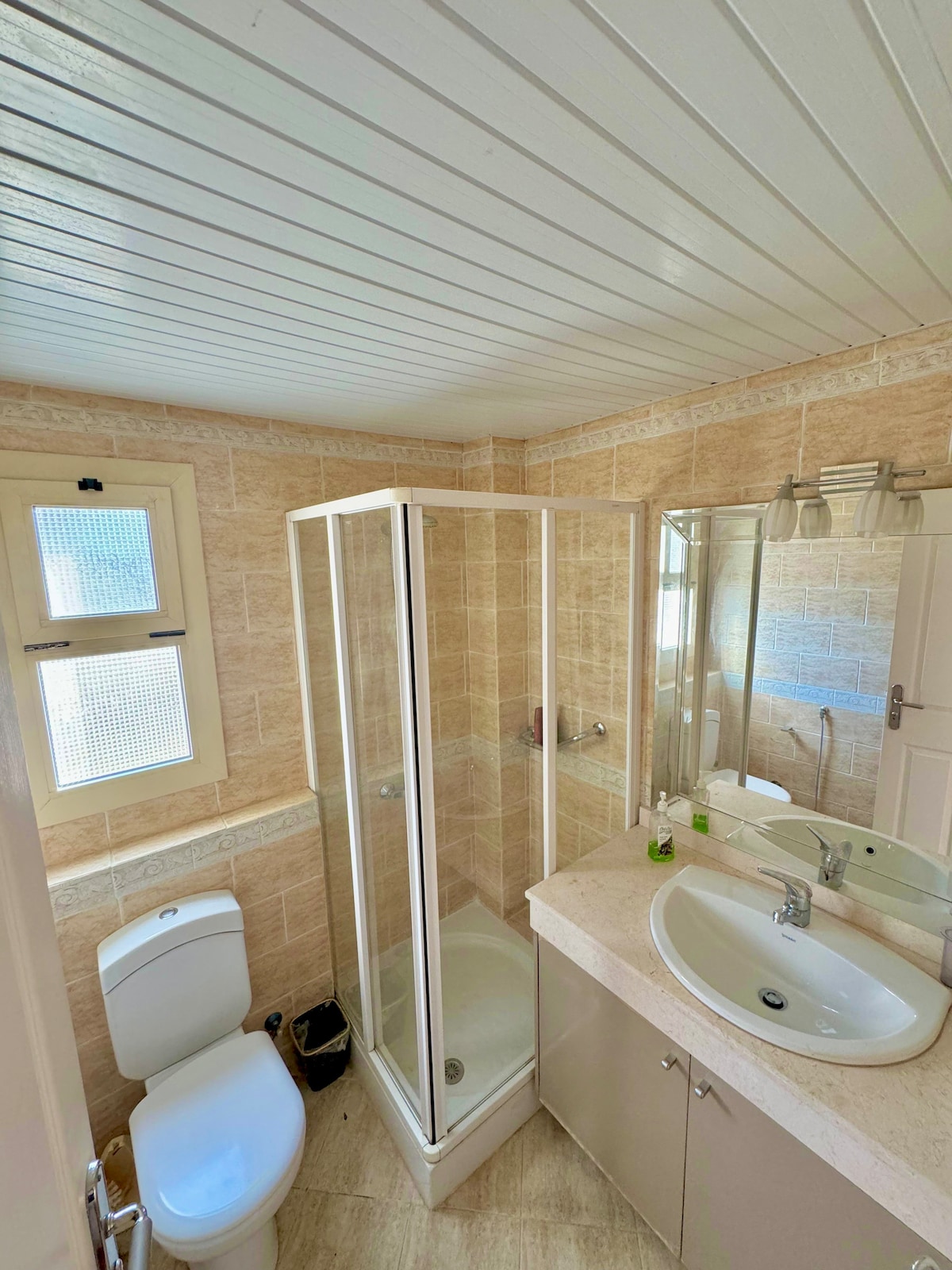 A well-appointed bathroom featuring a glass shower enclosure and a sink with a mirror above. A modern toilet and light-toned cabinetry are visible. Natural light filters in through a window, illuminating the beige tiled walls.